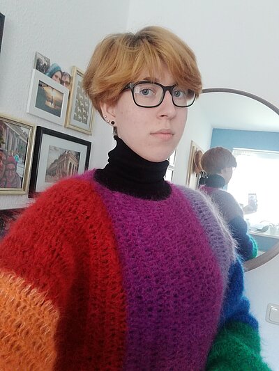 Michelle Michelle trägt kurze Haare, einen bunten Pullover und lächelt in die Kamera.