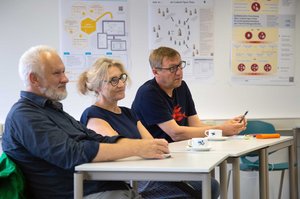 3 Personen sitzen am Tisch, im Hintergrund sind wissenschaftliche Poster