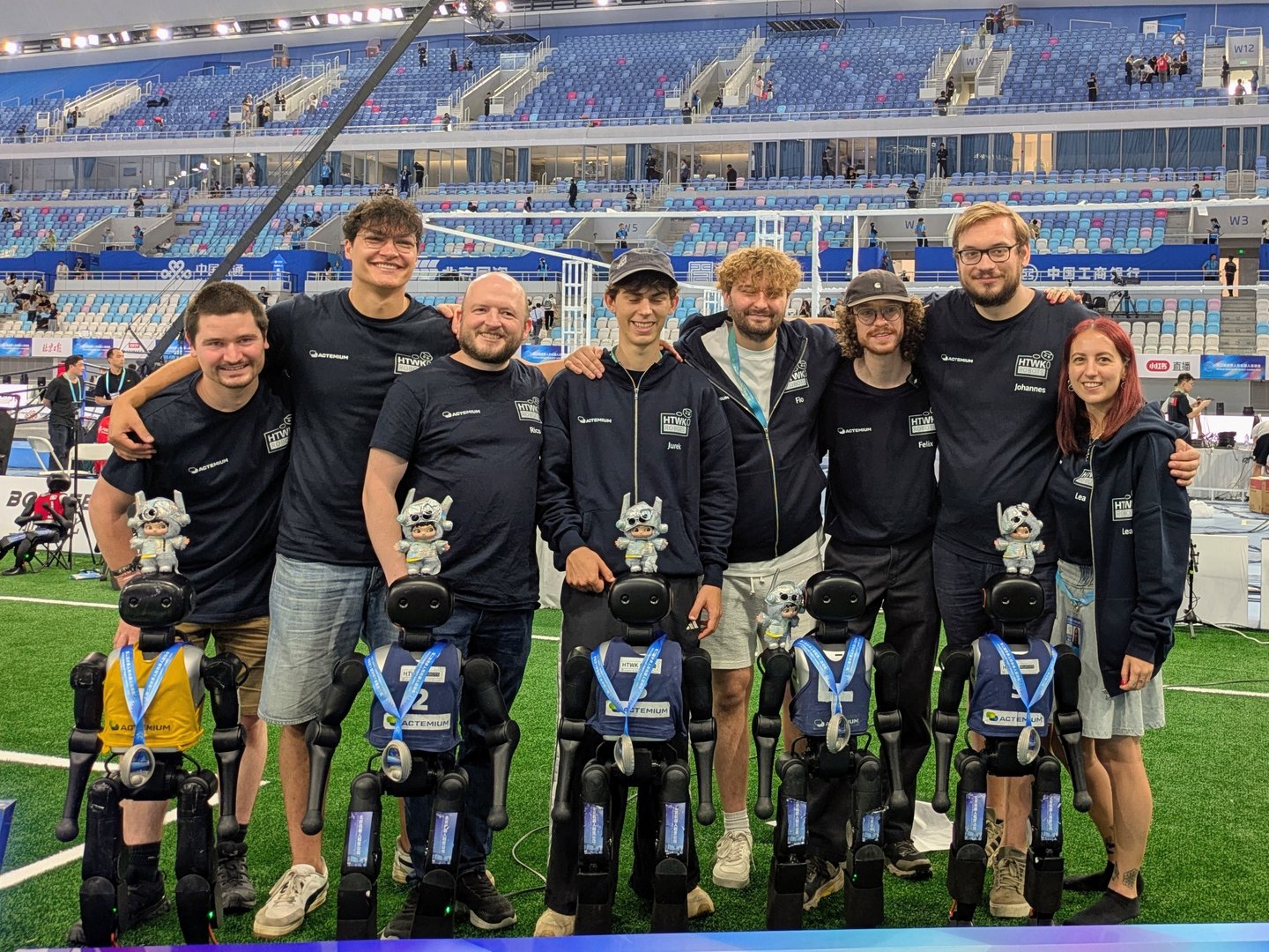 Die HTWK Robots nach den erfolgreichen Spielen in China. (Foto: HTWK Robots) Gruppenbild von Menschen und humanoiden Robotern in Stadion